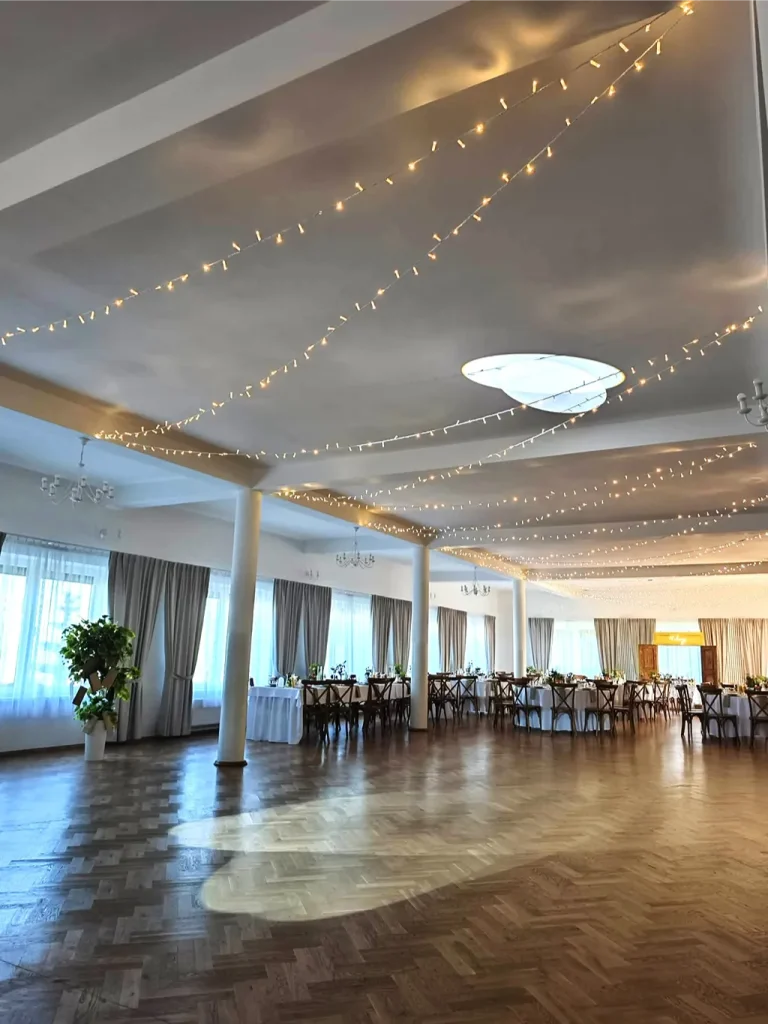 Duża sala bankietowa z drewnianymi podłogami, nakrytymi stołami i girlandami światełek zwisającymi z sufitu.